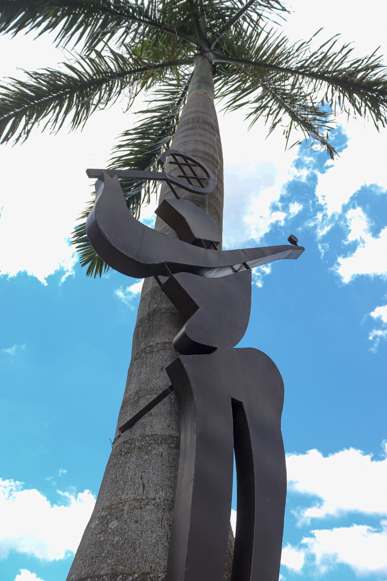 Escultura do boneco do Oeste Clube ao lado de uma palmeira, sob o céu azul